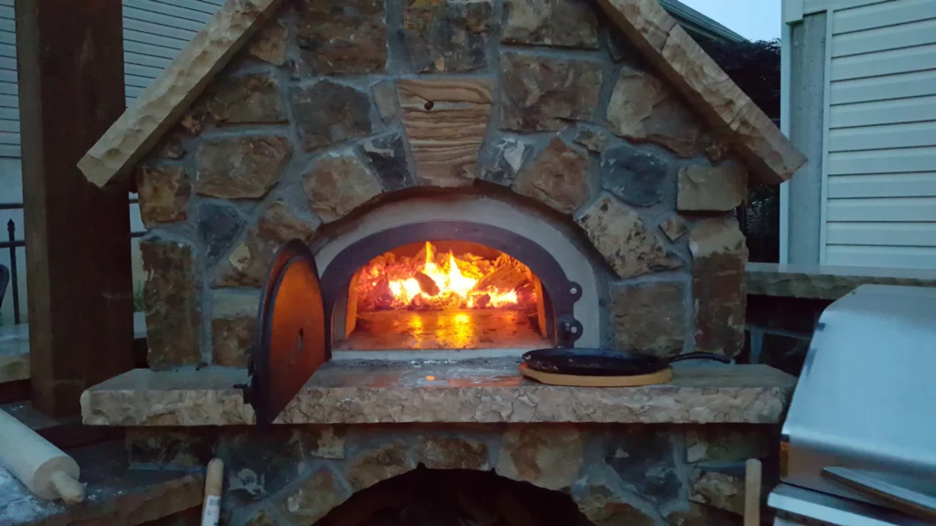 Full masonry outdoor pizza oven with fire lit and stone counter next to a stainless grill in Imperial, Missouri.